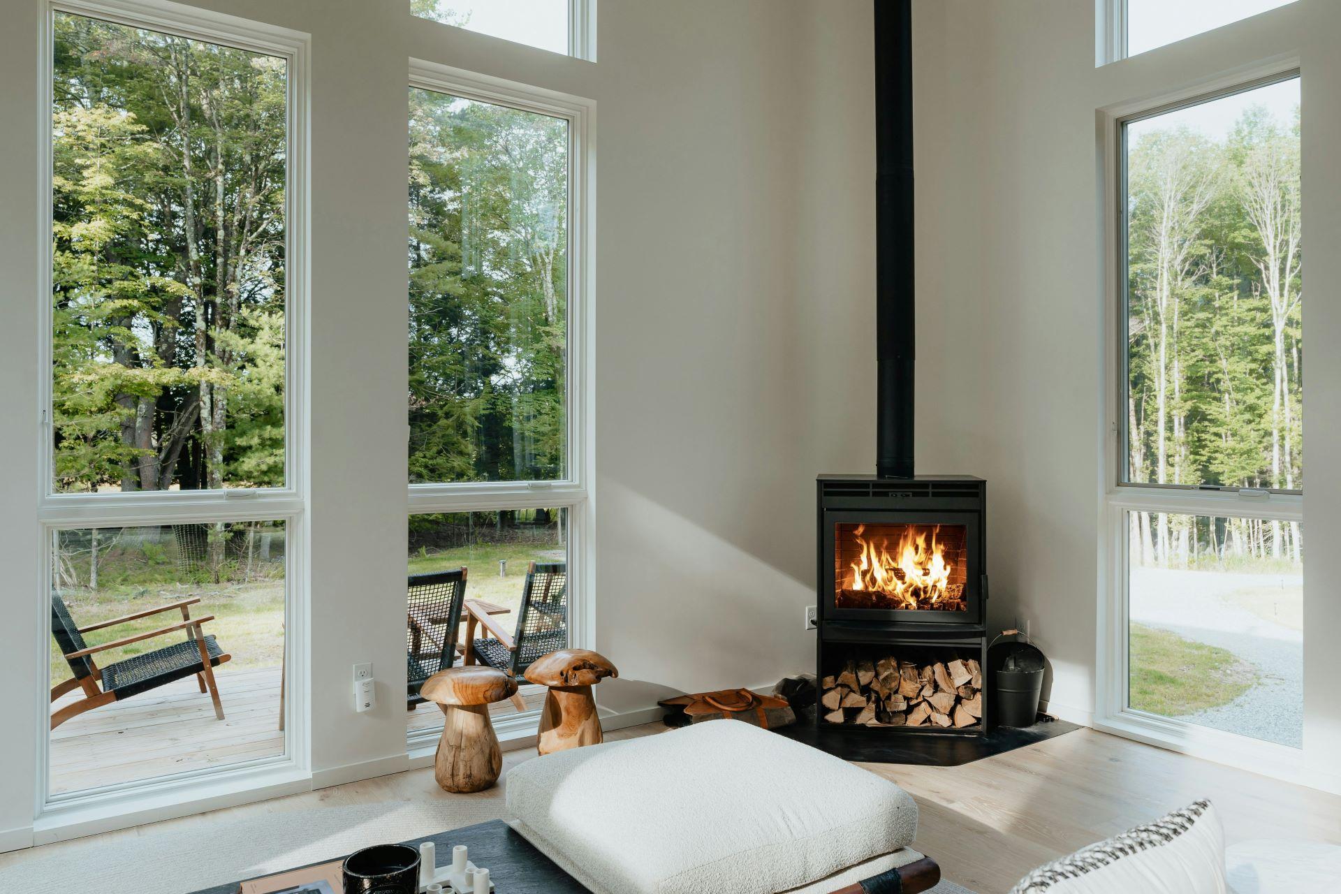 Stilvoller Wohnraum mit großen Fenstern, Kamin mit Glasscheibe und gestapeltem Holz darunter, umgeben von einer grünen Landschaft.